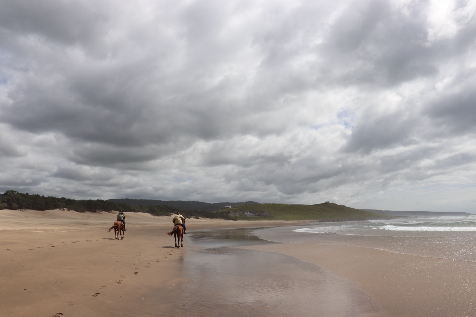 Two riders on horseback moving along the beach at Mbotyi under a wide cloudy sky
